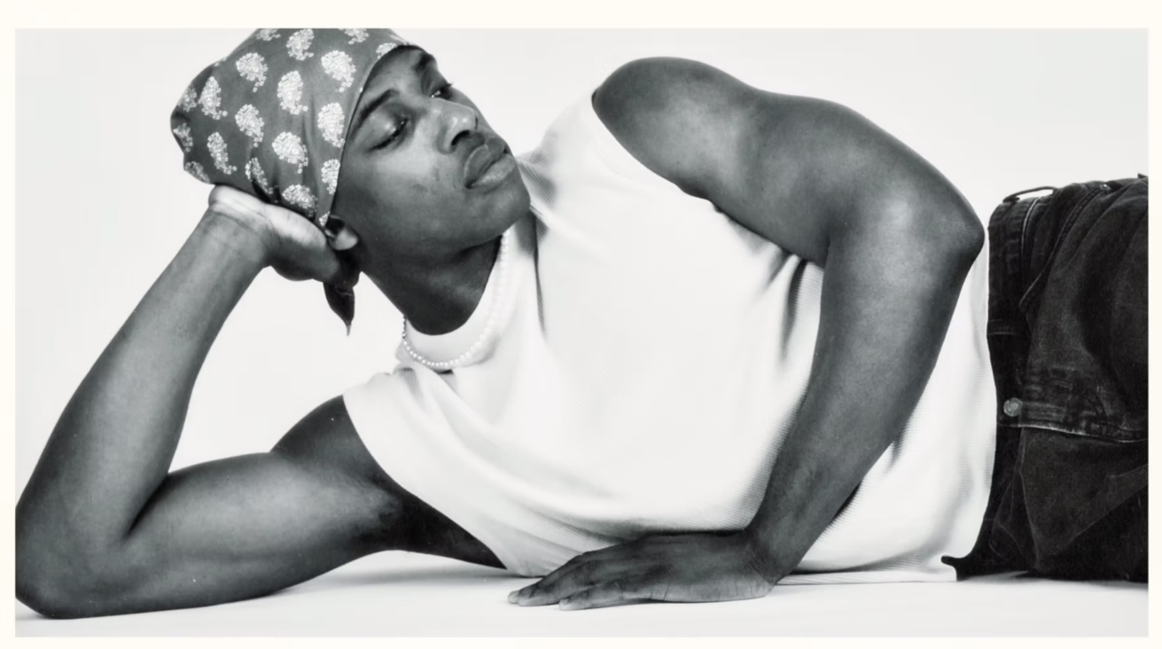 A person wearing a sleeveless shirt and bandana reclines on their side, propping their head up with one hand, looking off to the side against a plain background.
